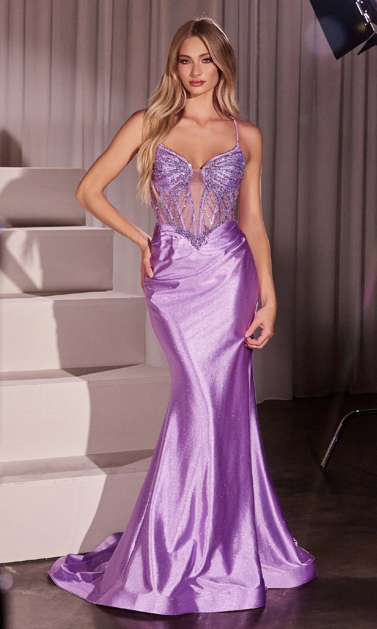 A woman stands indoors wearing the Ladivine CC8879 Beaded-Corset Long Prom Dress, featuring a shiny, fitted lavender design with a beaded, semi-sheer bodice and thin straps.